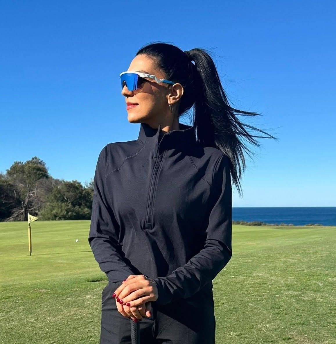 Rukmini Mehta in black golf attire and sunglasses standing on a clifftop green with the ocean behind
