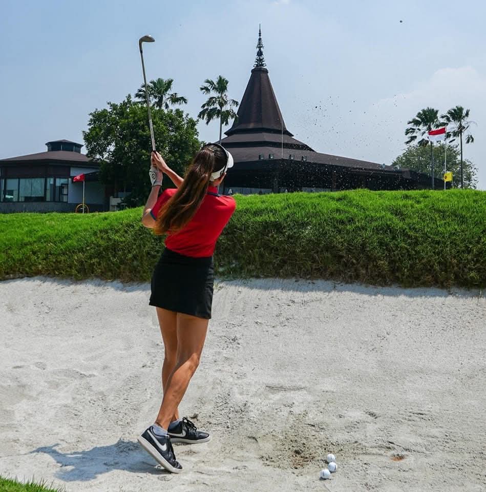 Rukmini Mehta playing a bunker shot with sand flying, pagoda-style clubhouse in the background