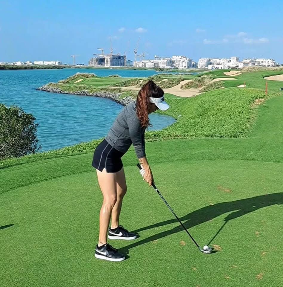 Rukmini Mehta addressing a golf ball on a waterside tee box at Yas Links with turquoise water behind