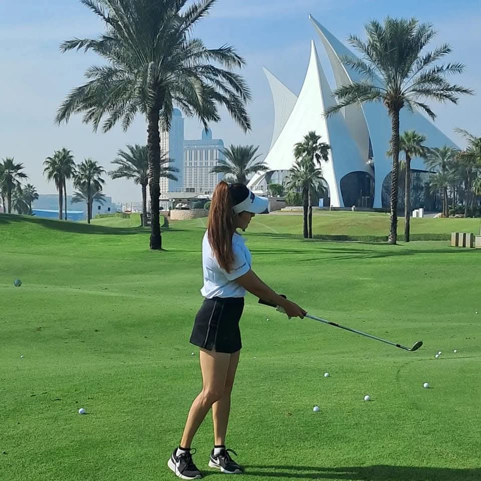 Rukmini Mehta chipping golf balls on a lush green with palm trees and a sail-shaped clubhouse behind