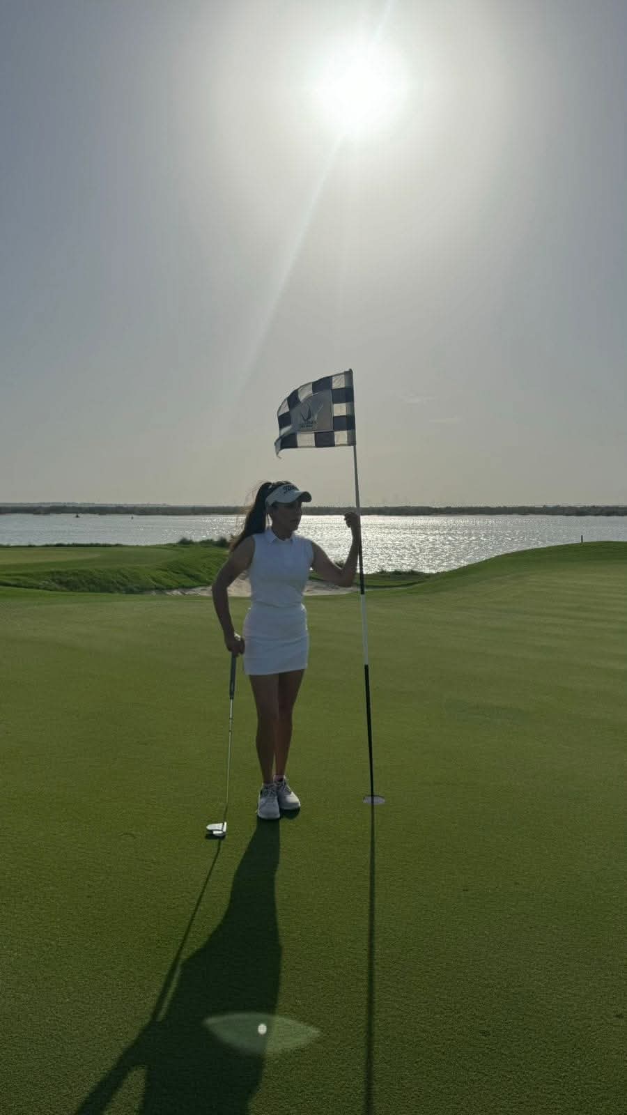 Rukmini Mehta holding the flagstick on a seaside green with sunlit water behind