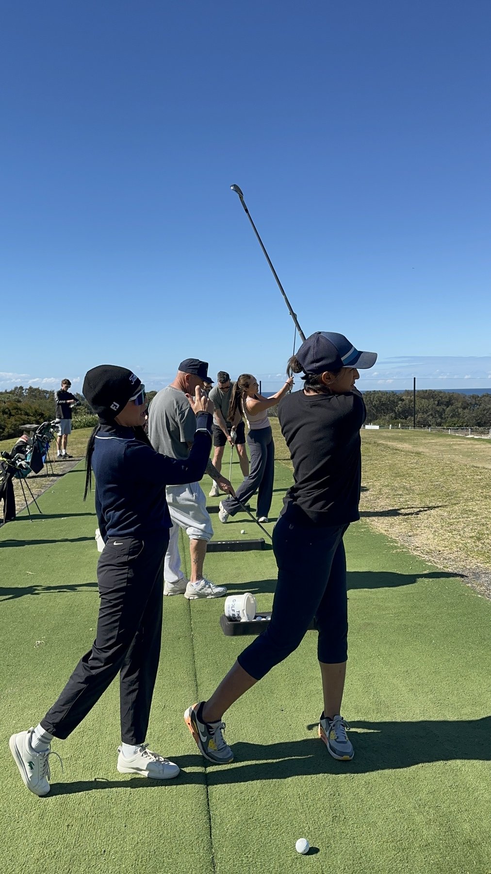 Rukmini Mehta coaching a golfer's iron swing at Cullens Range, St Michael's Golf Club, with the ocean on the horizon